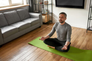 A calm man sitting on a green yoga mat in an apartment. This represents how medical nutrition therapy in Raleigh, NC can help you find relief from chronic conditions, disordered eating & more. Call us today to learn more. 