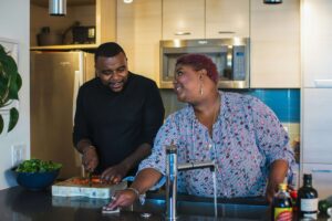 A couple cooking together in the kitchen & smiling at each other. Get started with our nutritional therapy in Raleigh, NC today by reaching out to us. We can help with disordered eating recovery. 