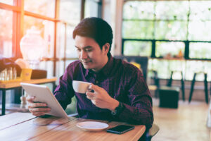 An asian man sitting down drinking coffee & reading a tablet. Medical nutrition therapy in Raleigh, NC can help with PCOS, IBS & more. Get started with a dietitian today.