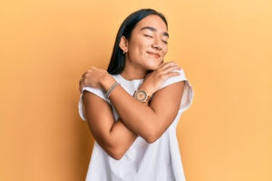 A young woman standing in front of an orange background hugging herself. Representing how nutritional counseling in Raleigh, NC can help enhance your self-care practices. Call now to begin working with a registered dietitian. 